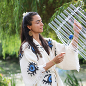 Eda Ertan playing the Quatrzophone (Crystal Harp) with a backdrop of running water and a calm willow tree faded in the corner. Eda is wearing the Nazar Kuftan by Soumak Boutique, with the protection blue eye as the main element of the design. 
